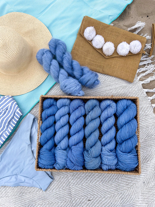 Blue yarn skeins in a basket with a straw hat, clutch, and towel on a textured surface.