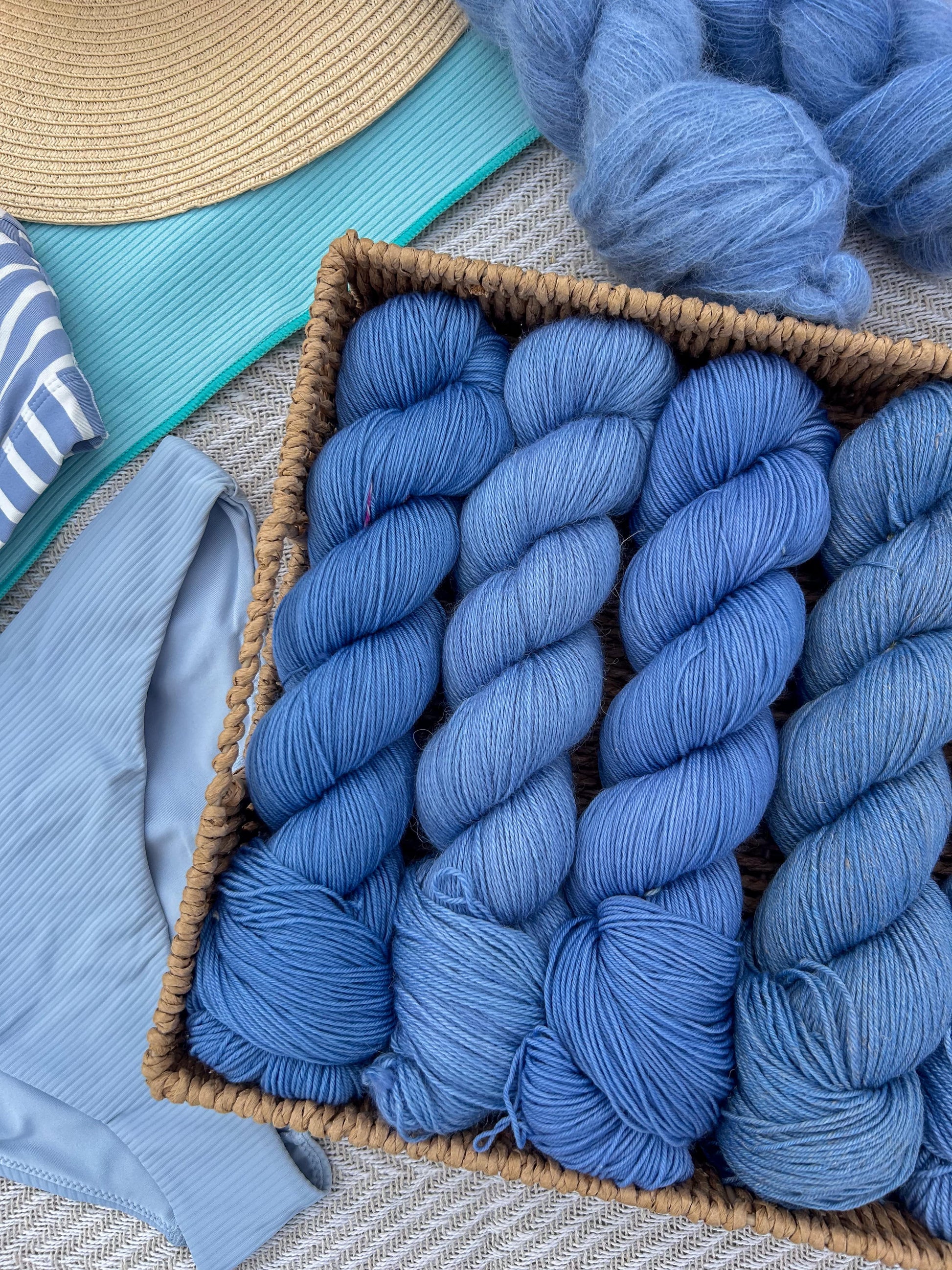 Blue yarn skeins in a basket with a straw hat and blue fabric in the background