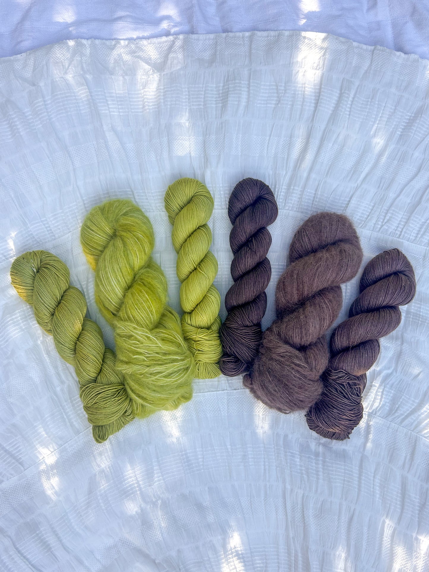 Three skeins of green yarn and three skeins of brown yarn on a textured white surface.